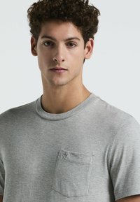 T-shirt en coton gris avec un col rond, des manches courtes et une poche poitrine gauche présentant un petit logo brodé. Texture lisse.