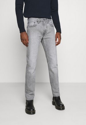 Mann trägt hellgraue Slim-Fit-Jeans, schwarze Stiefel und ein dunkles Langarmshirt, steht vor einem schlichten hellgrauen Hintergrund.