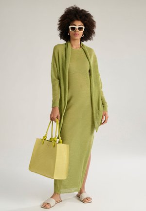 Green knit maxi dress with long sleeves, open front cardigan, and pale yellow handbag with knotted handles. Flat slide sandals are worn.