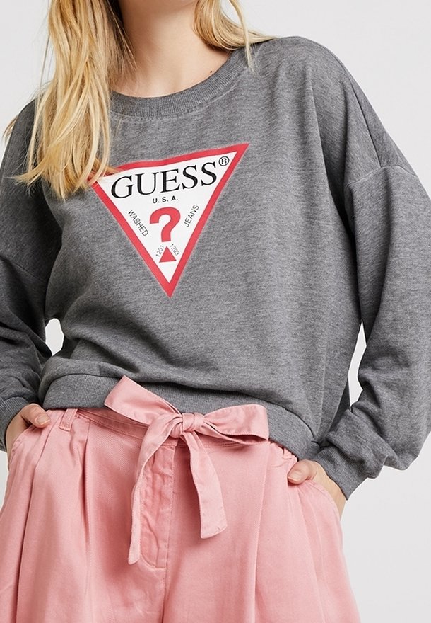 Femme portant un sweat-shirt gris Guess avec un logo rouge et blanc et un pantalon rose taille haute noué avec une ceinture en tissu.