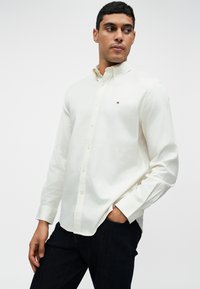 Camisa branca de botão em tecido liso, com um pequeno logótipo no peito, combinada com calças de ganga escuras.