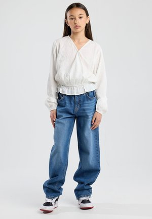 Weißes, strukturiertes Langarmshirt mit gerafftem Saum, kombiniert mit hellblauen, lässigen Jeans und schwarz-weißen Sneakern.