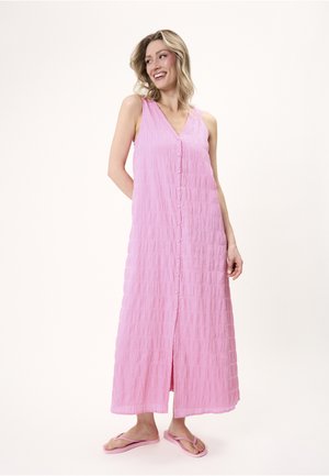 Lächelnde Frau trägt ein ärmelloses, knöchellanges pinkes Kleid mit Knöpfen vorne und pinke Flip-Flops, steht vor einem schlichten Hintergrund.