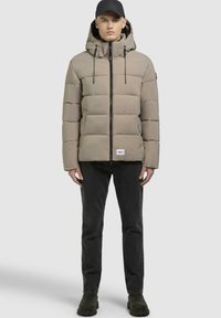 Polstret beige jakke med hætte, frontlynlås og snøre. Kombineret med sorte jeans og mørkegrønne ankelstøvler. Enkel design.