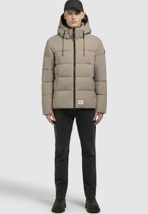 Padded beige jacket with a hood, front zipper, and drawstrings. Paired with black jeans and dark green ankle boots. Simple design.