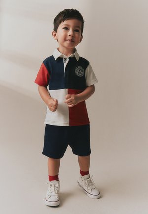 Ung dreng står iført en rød, hvid og navy bluse, navy shorts, røde sokker og hvide sneakers mod en ensfarvet beige baggrund.
