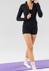 Schwarze sportliche Zip-Jacke mit langen Ärmeln, kombiniert mit schwarzen Shorts, weißen gestreiften Socken, auf einer lila Yogamatte, vor einem neutralen Hintergrund.