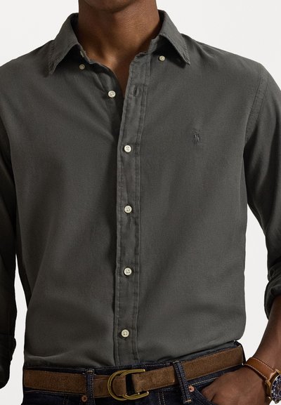 Tumši pelēks krekls ar pogām, ar teksturētu audumu, ar apkakli, baltām pogām un nelielu logo uz krūtīm. Nēsāts ar brūnu jostu.