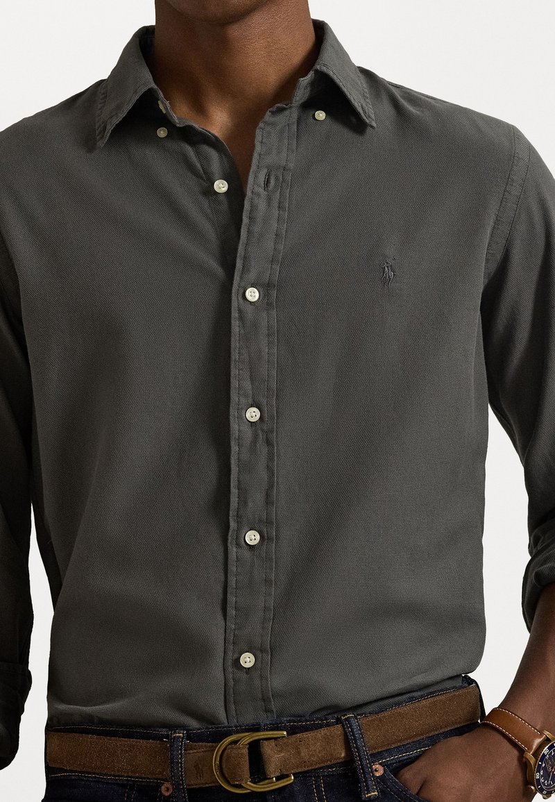 Dunkelgraues Hemd mit Knopfleiste aus strukturiertem Stoff, ausgestattet mit einem Kragen, weißen Knöpfen und einem kleinen Logo auf der Brust. Getragen mit einem braunen Gürtel.