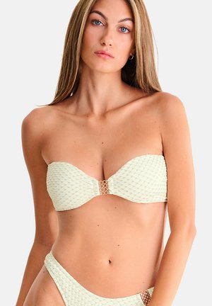 Femme aux longs cheveux raides portant un bikini crème texturé sans bretelles avec des connecteurs circulaires dorés sur le haut et le bas, sur fond blanc.