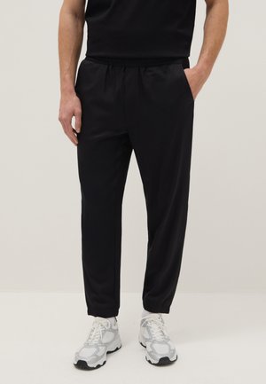 Man wearing black elastic waist pants with side pockets and white sneakers, standing against a plain background.