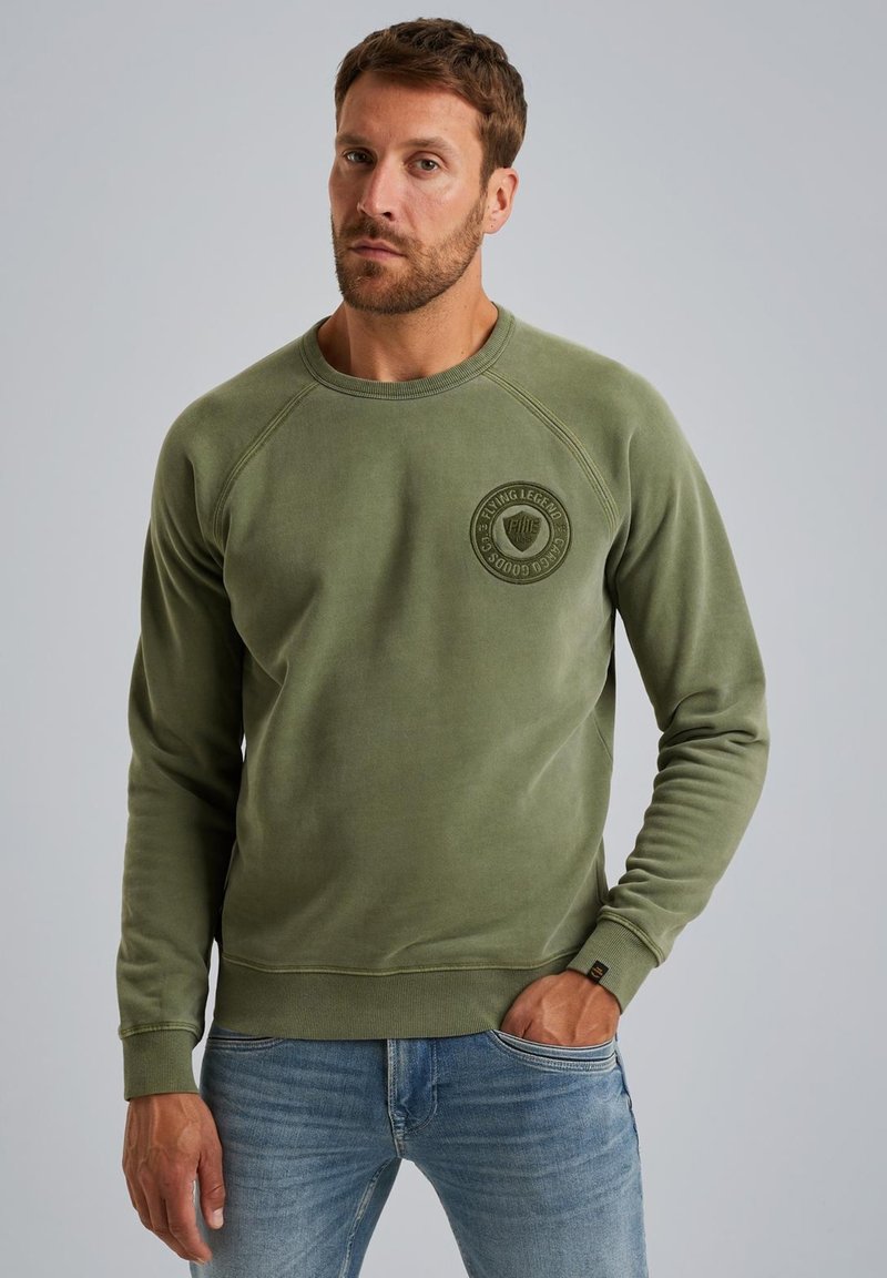 Olive green sweatshirt with a round neckline and ribbed cuffs, featuring a circular logo patch on the left chest. Smooth texture.