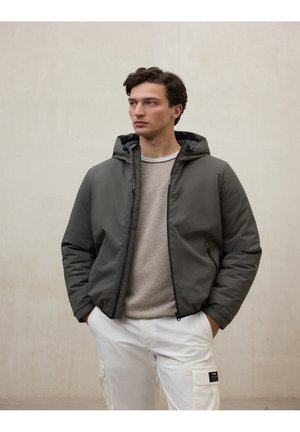 Hombre con chaqueta gris con capucha sobre suéter beige y pantalones cargos blancos, de pie con las manos en los bolsillos frente a un fondo liso.