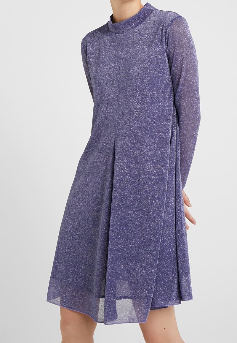Robe violette à manches longues jusqu'aux genoux avec un col montant et un subtil éclat portée par une personne debout devant un fond uni.