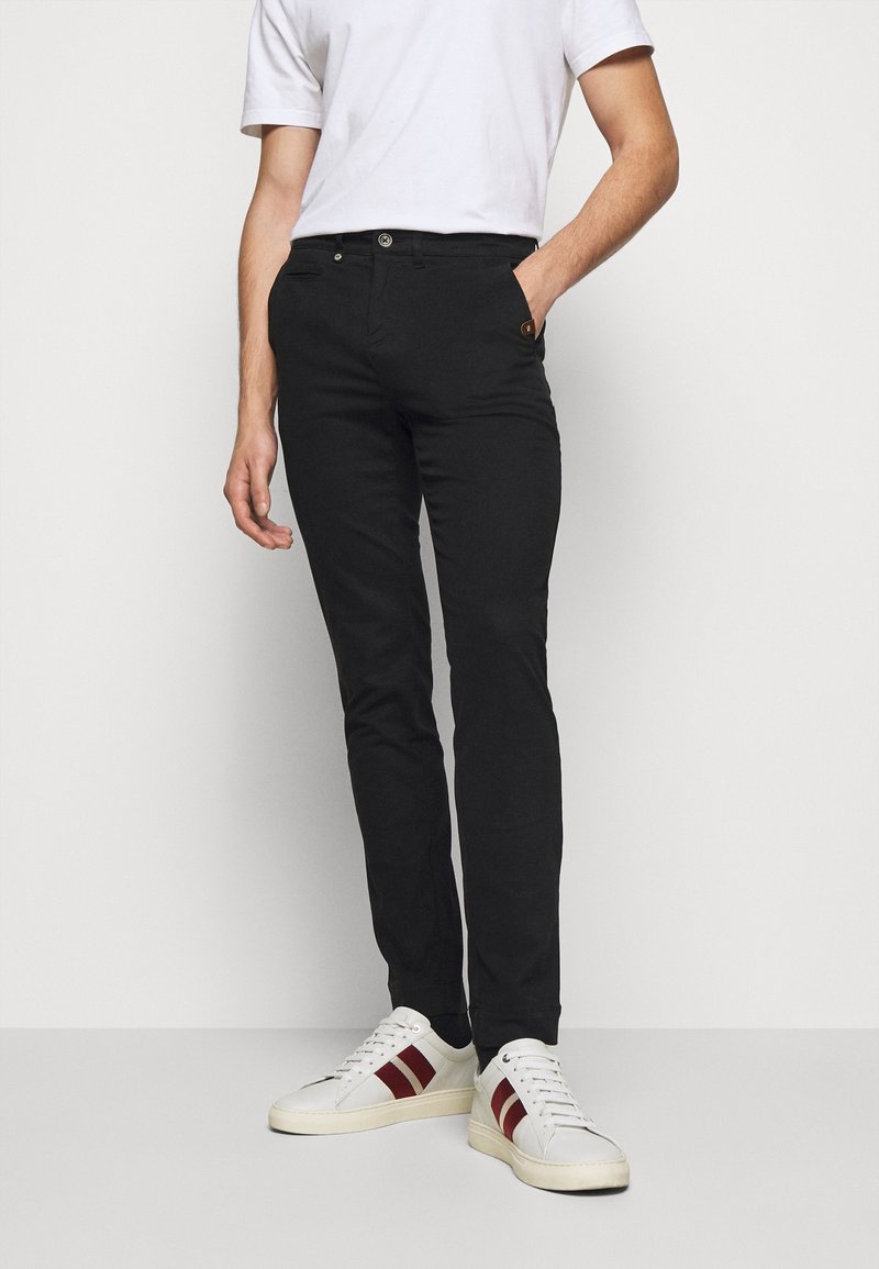 Homme portant un pantalon noir ajusté, un t-shirt blanc et des baskets blanches à rayures rouges, debout avec une main dans la poche.