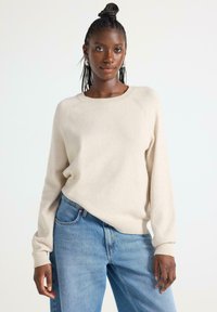 Vaalean beige neule, jossa on pyöreä pääntie ja joustinneulekäsiteet, yhdistettynä vaaleansinisiin farkkuihin. Neuleessa on rento malli ja pehmeä rakenne.