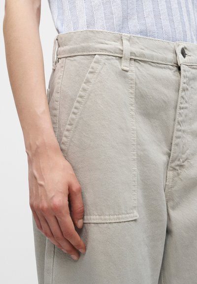 Main reposant à côté d’un pantalon beige clair avec poche avant visible et passants de ceinture, porté avec un haut blanc texturé.