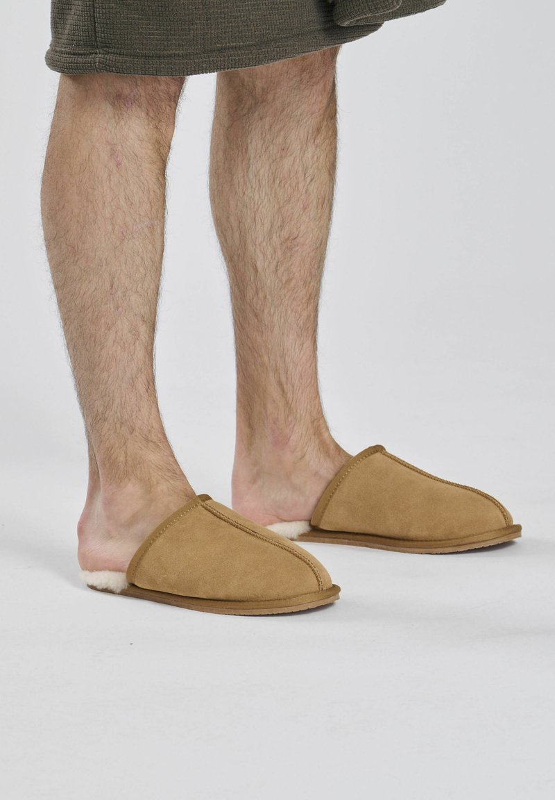 Bordeaux suède pantoffels met een zachte witte voering, afgerond ontwerp en een platte zool, weergegeven op blote voeten tegen een neutrale achtergrond.