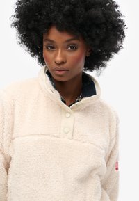 Cream-colored fleece pullover with a soft texture, half-button collar, and contrasting fabric accents. Features a red logo on the sleeve.