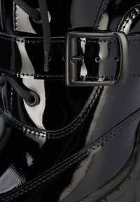 Černá lakovaná kožená bota s lesklým povrchem, kulatými šněrovadly a kovovou sponou. Prošívané detaily zdůrazňují design.
