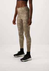 Siluetė tinkamos fizinės būklės asmens, vilkinčio smėlio spalvos maskuojančias legging'ų kelnes, juodas kojines ir juodus „Nike“ bėgimo batelius, prieš neutralų baltą foną.