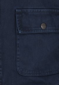 Marineblauer strukturierten Stoff mit einer Brusttasche, die mit einem silbernen Knopfverschluss versehen ist. Das Material weist ein diagonales Rippmuster auf.