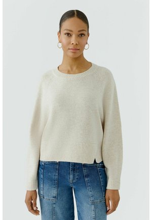 Femme aux cheveux attachés porte un pull en maille crème avec poignets et ourlet côtelés, associé à un jean bleu en denim avec des poches plaquées à l'avant.