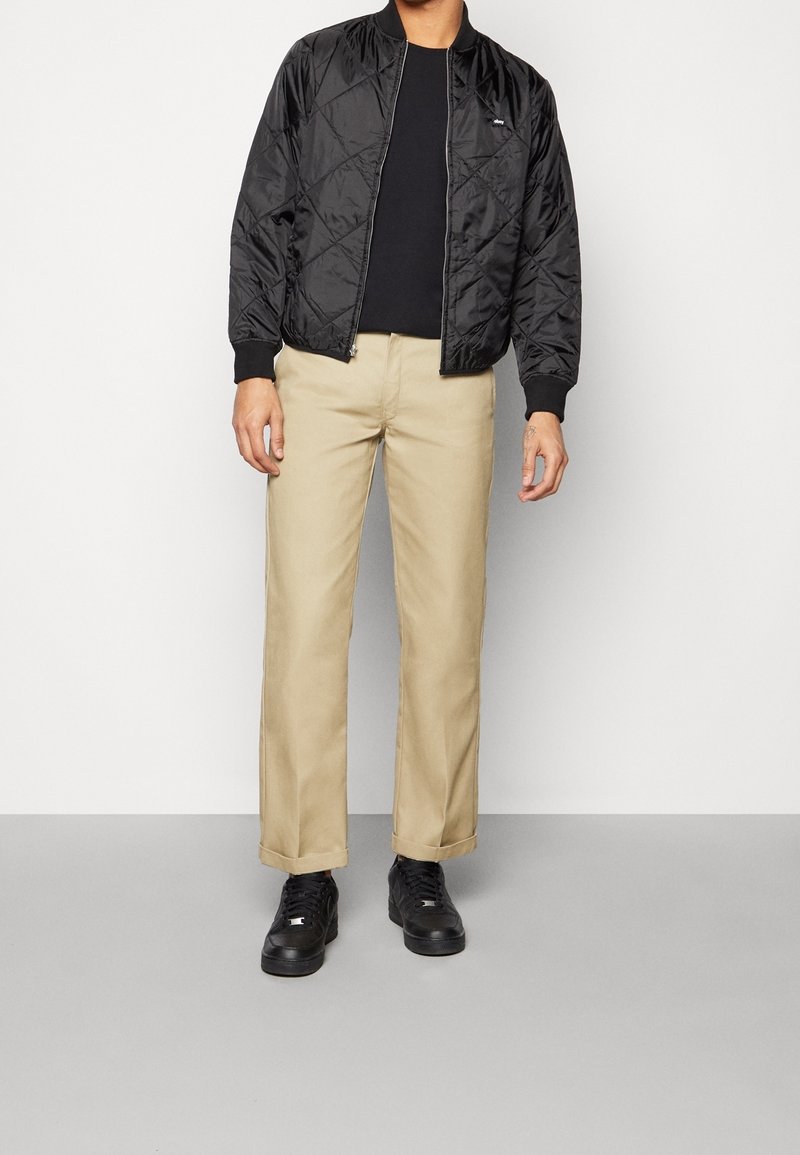 Veste bomber matelassée noire, pantalon beige à jambes droites, et baskets noires. Design simple avec poignets côtelés et fermeture éclair.
