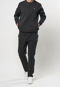 Muž oblečený v černé mikině a černých sportovních kalhotách, s bílými teniskami, stojící s rukama v kapsách.