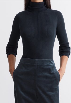 Vrouw draagt een zwarte geribbelde coltrui en donkerblauwe ribfluwelen broek met handen in de zakken tegen een effen achtergrond.