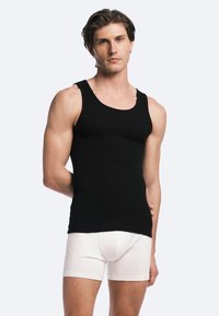 Schwarzes geripptes Tanktop mit breiten Trägern und Rundhalsausschnitt, kombiniert mit weißen eng anliegenden Boxershorts. Das Modell posiert frontal.