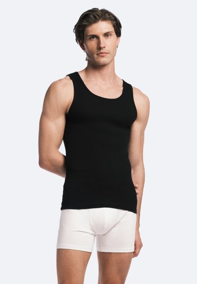 Débardeur noir à côtes avec des bretelles larges et un décolleté arrondi, associé à un boxer blanc ajusté. Le modèle pose de face.