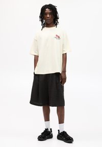 Světlě žluté př oversized tričko s grafickým potiskem; texturované černé šortky; černé tenisky; bílé ponožky. Jednoduchý, ležérní outfit.