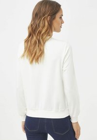 Sudadera blanca de corte relajado, con puños y dobladillo de canalé; tejido suave; llevada con vaqueros azul oscuro, mostrando un diseño simple y limpio.