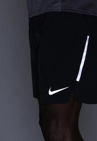 Schwarze Sportshorts mit reflektierendem weißen Nike-Logo und Akzentstreifen. Leichter Stoff mit leichtem Glanz und seitlichem Schlitz für Beweglichkeit.