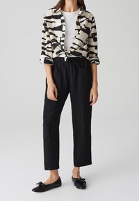 Chemise à boutons noirs et crème avec un motif abstrait, nouée à la taille ; associée à un pantalon noir à taille élastique et des chaussures noires plates.