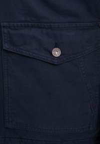 Marineblauwe canvas jas met een borstzak met een zilveren knop en klepsluiting, en een geweven stofdetail.