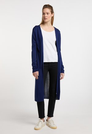 Langer navy-blauer Cardigan aus weichem Material, mit Kapuze, offenem Vorderteil und ribbelten Bündchen, getragen über einem weißen Oberteil und schwarzen Jeans.