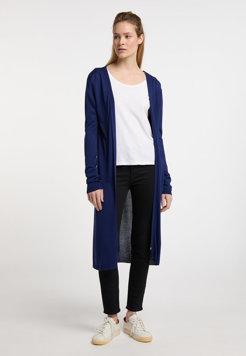 Lang navy cardigan lavet af blødt stof, med hætte, åben front og ribbede manchetter, båret over en hvid top og sorte jeans.