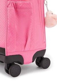 Rosa hårdskalresväska med ett strukturerat skalfmönster, utrustad med svarta hjul och en sidoficka. Fäst vid väskan finns en fluffig rosa pompongcharms.