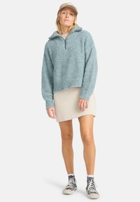Pullover peloso color teal con colletto a mezza zip, mini gonna beige a costine e sneakers grigie, abbinate a calze bianche e un cappellino beige.