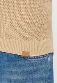 Pull en tricot beige rentré dans un jean en denim bleu, montrant un motif tricoté texturé et une petite étiquette marron à l'ourlet.
