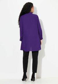 Fialová tunika s dlouhým rukávem a límečkem, s rozparkem na zadní straně a zapínáním na knoflíky na manžetách, spárovaná s černými těsnými kalhotami a černými kotníkovými botami.