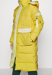 Keltainen toppatakki tikkikuviolla, valkoiset kontrastiyksityiskohdat, huppu, suuri etutasku ja Jordan-logo hihaosassa.