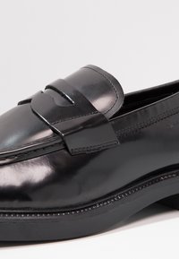Sapato de couro preto com acabamento brilhante, apresentando um bico arredondado, realces de tiras proeminentes e uma sola de borracha elegante para maior aderência.