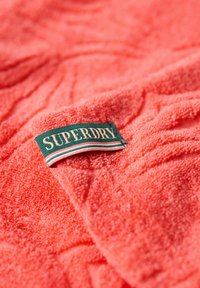 Koralowy ręcznik frotte o teksturowanej powierzchni, z zieloną metką z napisem „SUPERDRY” oraz białymi i czerwonymi paskami poniżej.