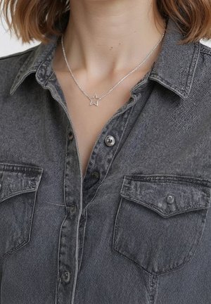 Femme portant un collier avec un pendentif en forme d'étoile argentée sur une chemise en jean gris foncé boutonnée avec des poches poitrine.