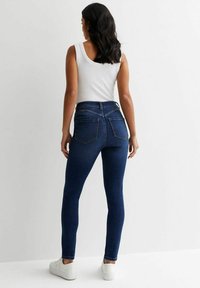 Jeans skinny azul oscuro con cintura alta y bolsillos traseros, combinados con una blusa sin mangas blanca y zapatillas blancas. Textura de denim suave.