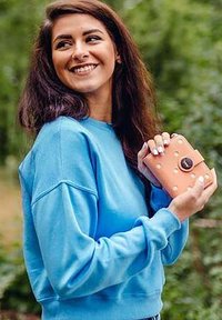 Růžová peněženka s bílými puntíky a černým kruhovým zapínáním, držena v ruce, spárována se světle modrou mikinou, venku v zeleném prostředí.