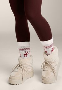 Bézs színű, bolyhos bokacsizma vastag talppal, fehér zoknival, amelyen piros rénszarvas és hópihe minták láthatók, valamint bordó leggings. 
